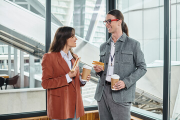 young coworkers having sandwich with tea on break and smiling sincerely at each other, coworking