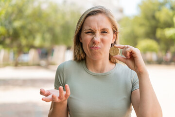Young blonde woman holding a Bitcoin at outdoors making doubts gesture while lifting the shoulders