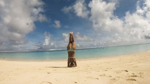 Asian tanned skin female girl woman doing Yoga inversions pose at a white sandy beach and shallow calm lagoon of turquoise blue water during daytime sunshine weathe in Aroa Rarotonga Cook Islands