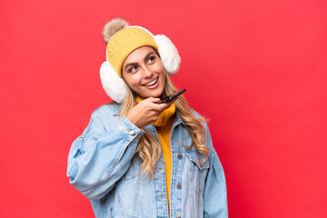Young pretty Uruguayan woman wearing winter muffs isolated on red background background keeping a...