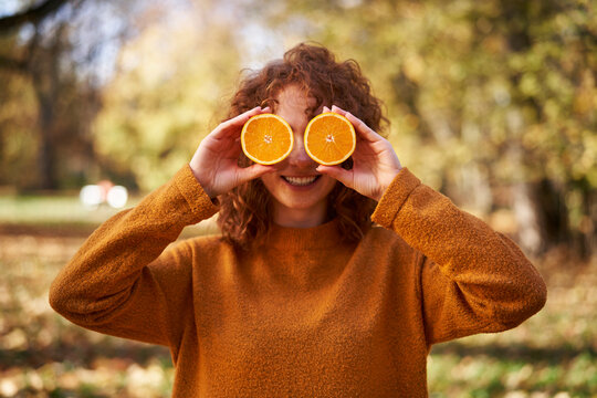 Smiling redhead woman holding orange slices in front of face at autumn park