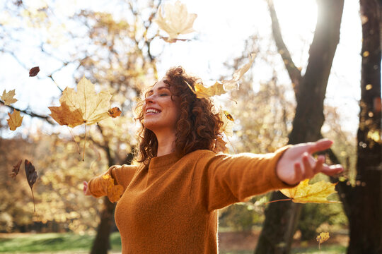Autumn leaves falling on redhead woman with arms outstretched at park