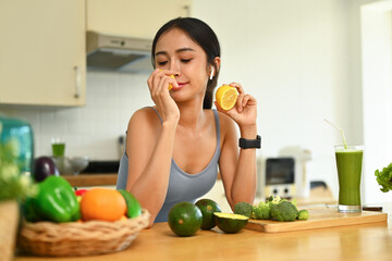 Beautiful young woman smelling fresh lemon with closed eyes while preparing ingredients for making healthy food in kitchen