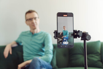 Unfocused man is sitting on sofa in background, focusing on phone on tripod with picture of camera shooting. Blogger.