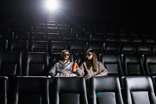 A couple in love is watching a movie with 3D glasses and drinking from a red cup. Cinema for two