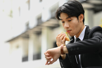 Busy asian male office worker in a hurry biting sandwiches and checking time on wristwatch for appointment or transportation