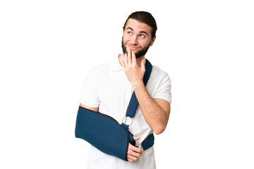 Young handsome man with broken arm and wearing a sling over isolated chroma key background looking...