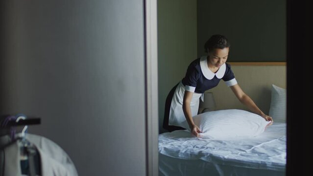 Medium wide shot of maid making bed and fluffing pillow in hotel room