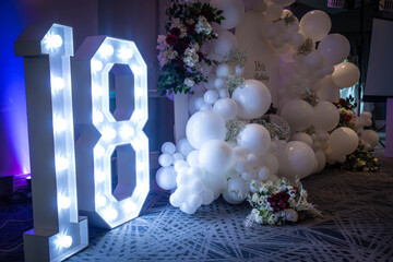 An 18 decorative sign lit up, balloons and a sign form a backdrop at an 18th birthday party
