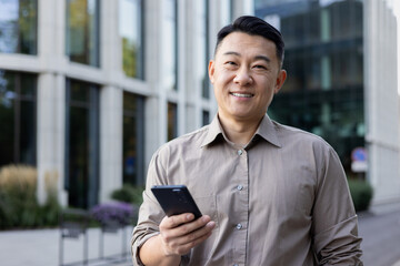 Portrait of Asian young man businessman, worker, manager standing outside office center and using mobile phone. Smiling at the camera. Close-up photo