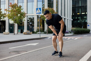 A young Asian man runs in the morning through the streets of the city, stands and holds his leg,...