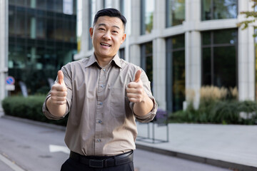 Portrait of a successful and confident Asian male businessman standing near an office center, smiling at the camera and showing the super sign with his fingers