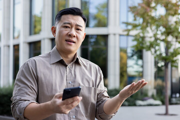 Close-up portrait of a sad young Asian man standing near an office building with a phone in his...