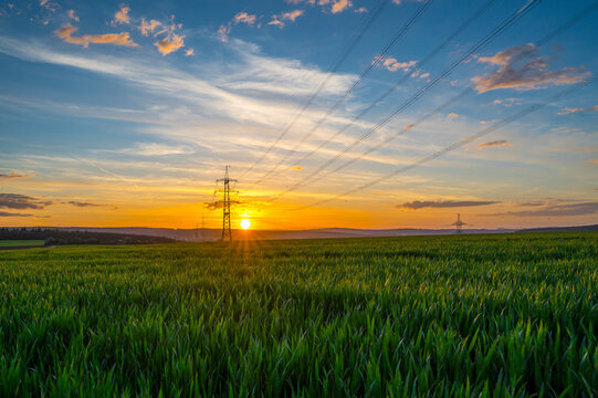 Germany, Hesse, Hunfelden, Electricity pylon in vast green field at summer sunset
