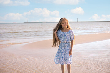 little girl in a dress on the beach with developing long blond hair in the wind and looking on the sea.