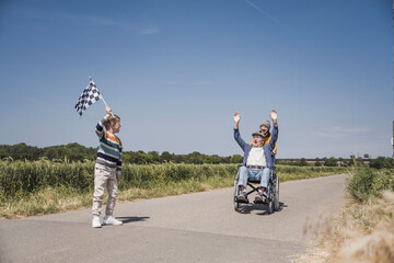 Boy holding checkered flag with senior man sitting in wheelchair on road