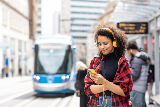 Smiling woman wearing headphones and waiting for tram in city