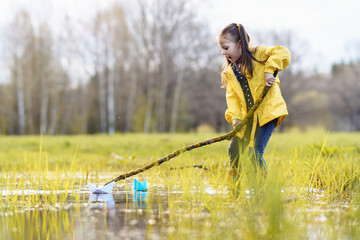 Amazed kid standing in puddle on lawn and looking at paper boats with open mouth, playing with ships using branch.