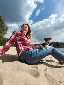 A young beautiful woman sits on the sand on the river bank with a pet dog of the jack russell terrier breed in her arms