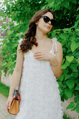 Portrait of a young woman walking on a summer day. A beautiful model in a white dress and sunglasses poses outside