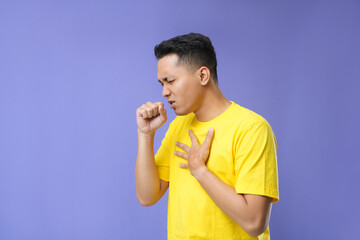 A young Asian man feeling a sore throat and flu symptoms, coughing with his mouth covered. Isolated on a purple background.