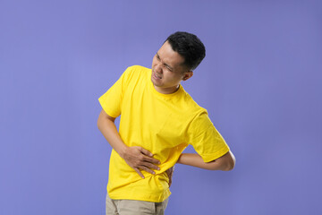 Shouting a young Asian man suffering from back pain isolated on a purple background. Health problem, holding him back.