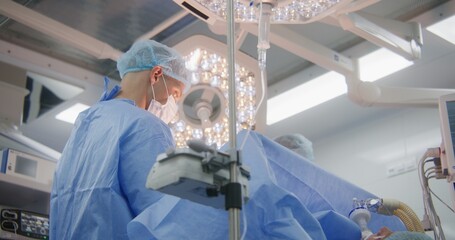 Professional adult surgeon in surgical suit operates patient with colleague and talks in operating...
