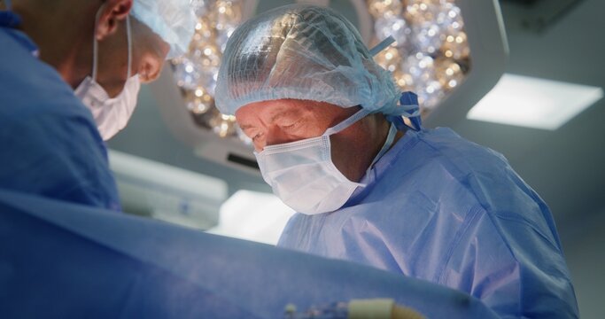 Professional surgeon in surgical suit with adult colleagues operates patient in modern operating room. Male medics cauterize cut. Patient lies on table under anesthesia with breathing tube.