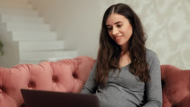 Beautiful young smiling woman working on laptop while sitting on sofa in living room at home