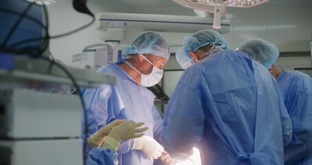 Young nurse prepares medical clip with thread and gives it to doctors. Professional surgeons in surgical suits stitch wound. Medics operate patient in surgery room. Medical staff work in hospital.