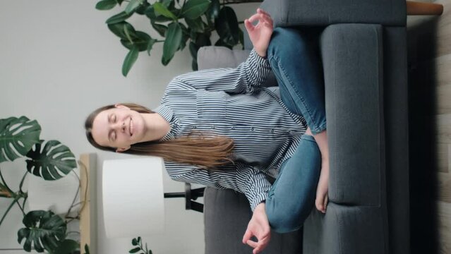 Vertical footage of cute young woman sits cross-legged on couch at home, doing meditation practice, boosting inner balance, improve self-conscious, mindfulness state, harmony and breathing exercises