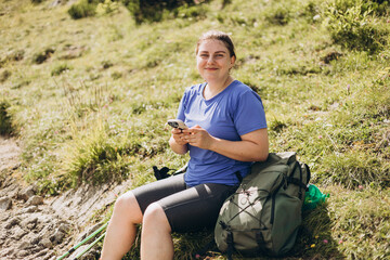 Naklejka na ściany i meble Stylish woman with green backpack holding smart phone and relaxing in nature. Hiking active female lifestyle siting outdoors.