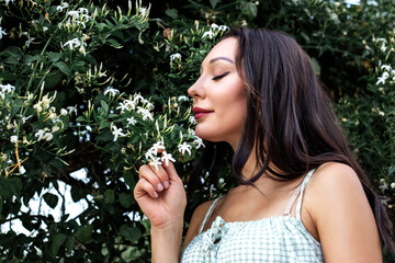 Portrait of a beautiful young caucasian brunette woman smelling jasmine flowers outdoors - obrazy, fototapety, plakaty