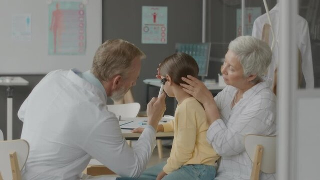 Waist up of mature Caucasian male otolaryngologist examining ears of little girl with special instrument during medical appointment in hospital