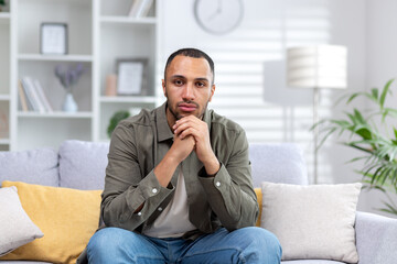 Fototapeta premium Portrait of a young hispanic man looking seriously at the camera, sitting at home on the couch with folded hands
