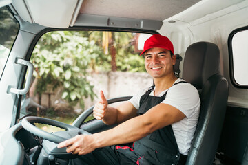 Obraz premium Happy professional truck driver with his assistant wearing a red cap thumb up, smiling, looking at the camera from a truck window before delivering parcel. A truck driver and delivery service concept.