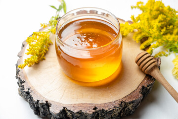 A jar of liquid honey on a light background close-up, next to flowers and a spoon for honey.