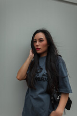 beautiful cool stylish Asian woman with red lips in a fashion urban outfit with a leather bag walks on a windy day in the city near a wall