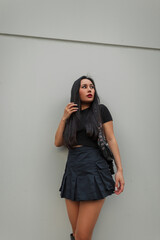 Fashionable beautiful sexy model Asian woman in fashion urban summer outfit with black t-shirt and skirt with trendy bag stands near the grey wall in the city
