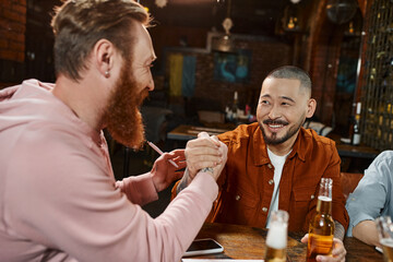 joyful asian man shaking hands with bearded colleague while closing deal near beer bottles in pub