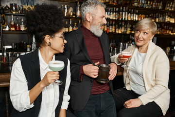 multiethnic women with cocktails glasses smiling during conversation with bearded colleague in bar