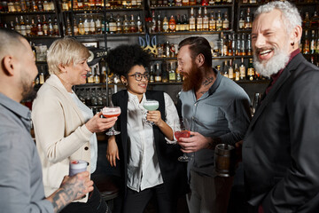 bearded tattooed man talking to smiling african american woman near workmates with drinks in bar