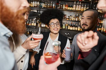 positive african american woman holding cocktail and smiling near multiethnic work friends in bar