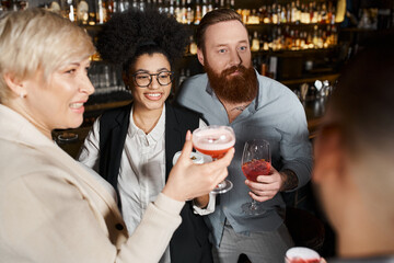 bearded tattooed man and african american woman smiling near workmates with cocktail glasses in bar