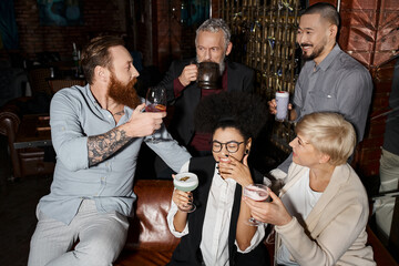 bearded tattooed man talking to happy multiethnic colleagues with drinks in bar, after work leisure