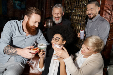 bearded tattooed man clinking glasses with multiethnic women near smiling workmates in cocktail bar