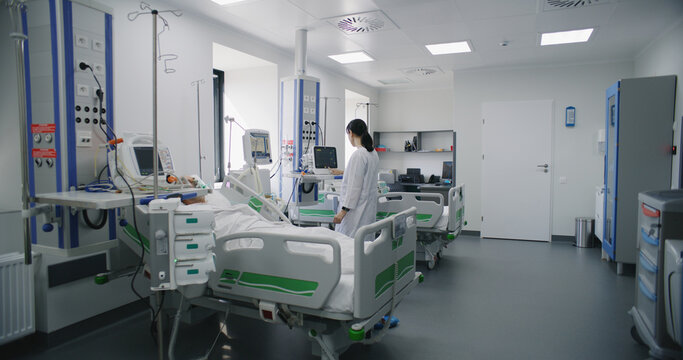 Emergency room with modern equipment in hospital. Elderly man in oxygen mask lies and sleeps in bed after successful surgery. Nurse works on computer in intensive care department of medical facility.