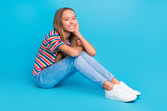 Full size photo of optimistic cute teen girl wear colorful t-shirt pants sit on floor hand on cheekbone isolated on blue color background