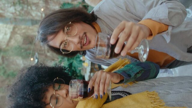 Cheerful Latin couple holding wine and talking on camera, making selfie or video calling during barbecue party in the backyard. POV shot, video portrait. Vertical format footage
