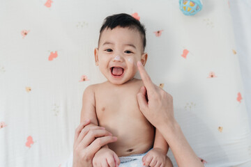 Happy Asian Baby with a Comfortable Smile, Mother Finger Gently Applying Skin Care Lotion on His...